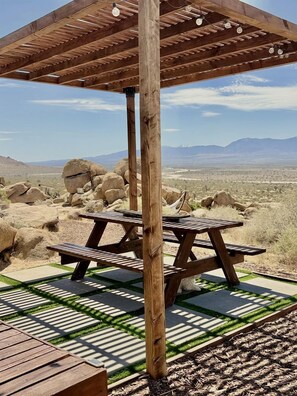 A wooden pergola with a picnic table, set against a desert landscape with mountains in the background. - Stunning Geo Dome with Joshua Tree vibes (Palmdale)