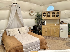 Un dormitorio con cama con dosel, un armario de madera, un refrigerador pequeño y una planta.