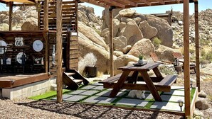 A wooden pergola with a picnic table and chairs, surrounded by rocks and desert vegetation. - Stunning Geo Dome with Joshua Tree vibes (Palmdale)
