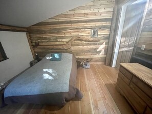 Un dormitorio con techo inclinado de madera, una cama, un aparador y un ventanal pequeño.