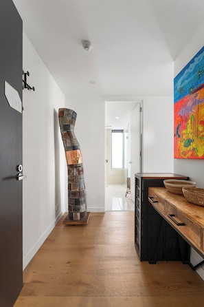 Ein Flur mit Holzboden, einer Metallskulptur, einer Holzbank mit Körben und einem farbenfrohen, abstrakten Gemälde an der Wand.