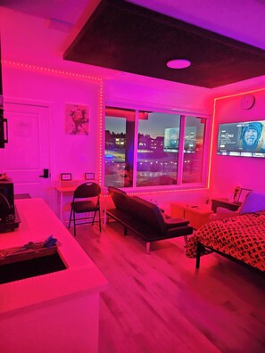 A room with a bed, a desk, a chair, a sofa, and a window with a city view. - Swithome Properties Cozy Studio Pond View, Near Yeg Airport (Edmonton)