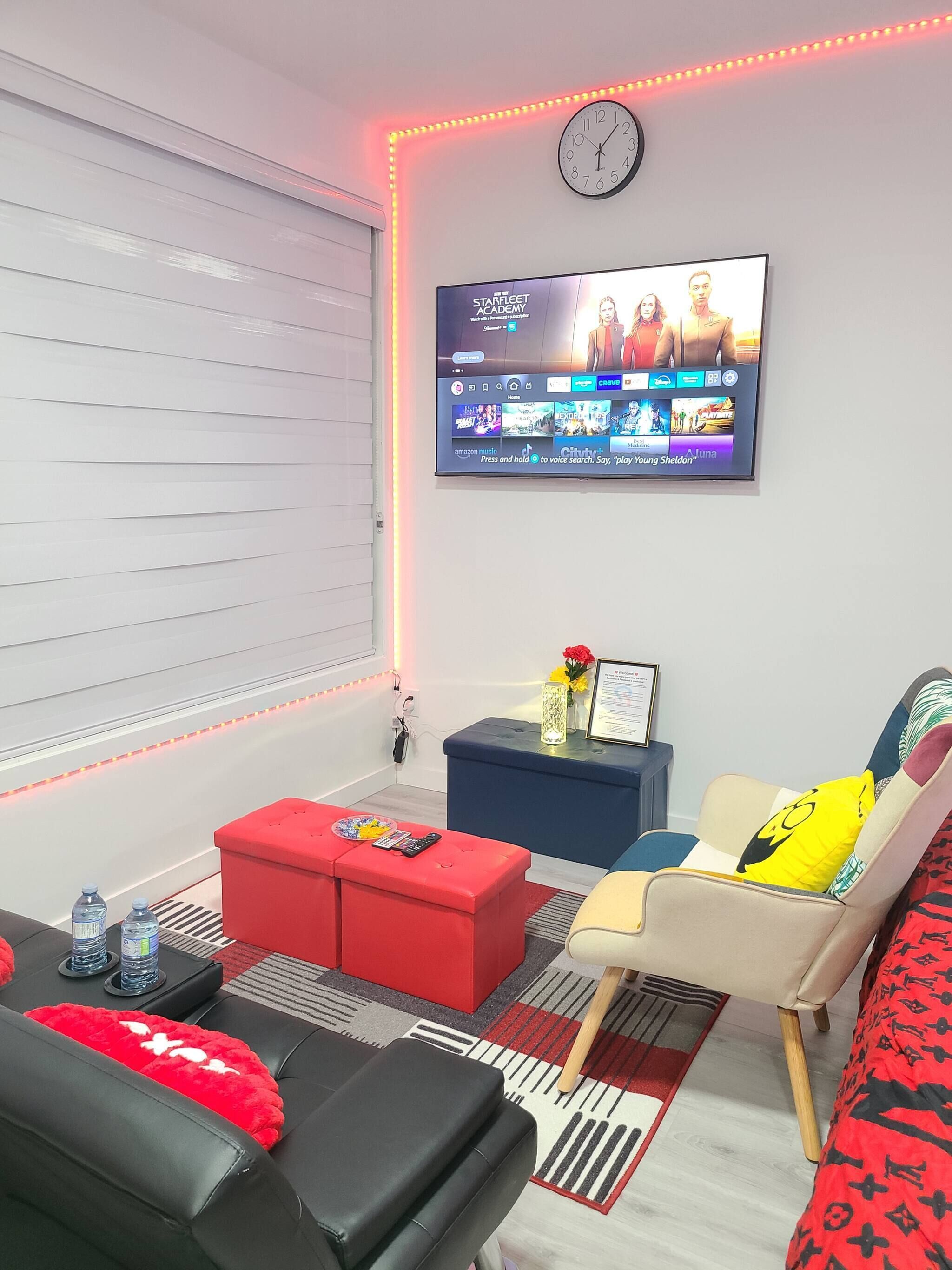 A modern living room with a flat-screen TV mounted on the wall, a red ottoman, a blue bench, and a beige armchair.