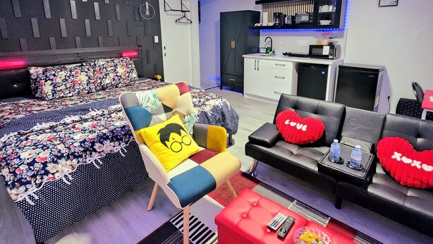 Ein modernes Schlafzimmer mit Bett, einer Couch, einem roten Hocker, einer Küchenzeile und einer Deckenleuchte.