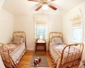 Une chambre avec deux lits simples, un ventilateur de plafond, une fenêtre avec des rideaux, une petite table avec une lampe et un tapis sur le sol.