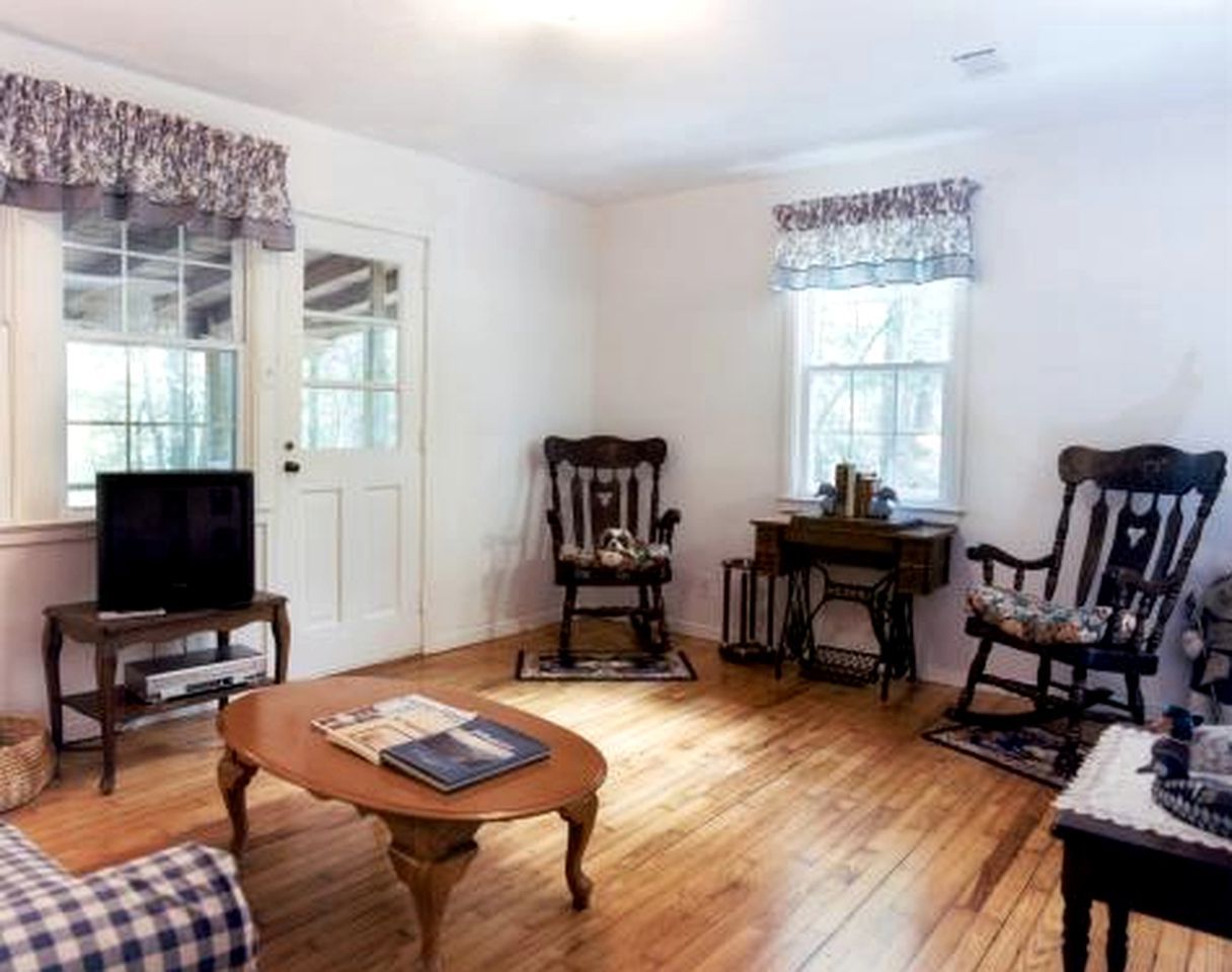 A living room with a television on a stand, a coffee table, two chairs, a small table with items, and a window with curtains.