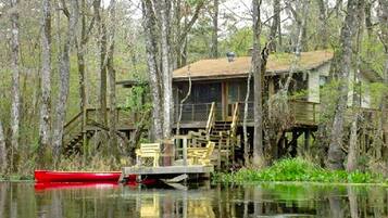 Une maison sur pilotis entourée d’arbres, un quai avec un bateau rouge et une terrasse en bois avec des chaises.