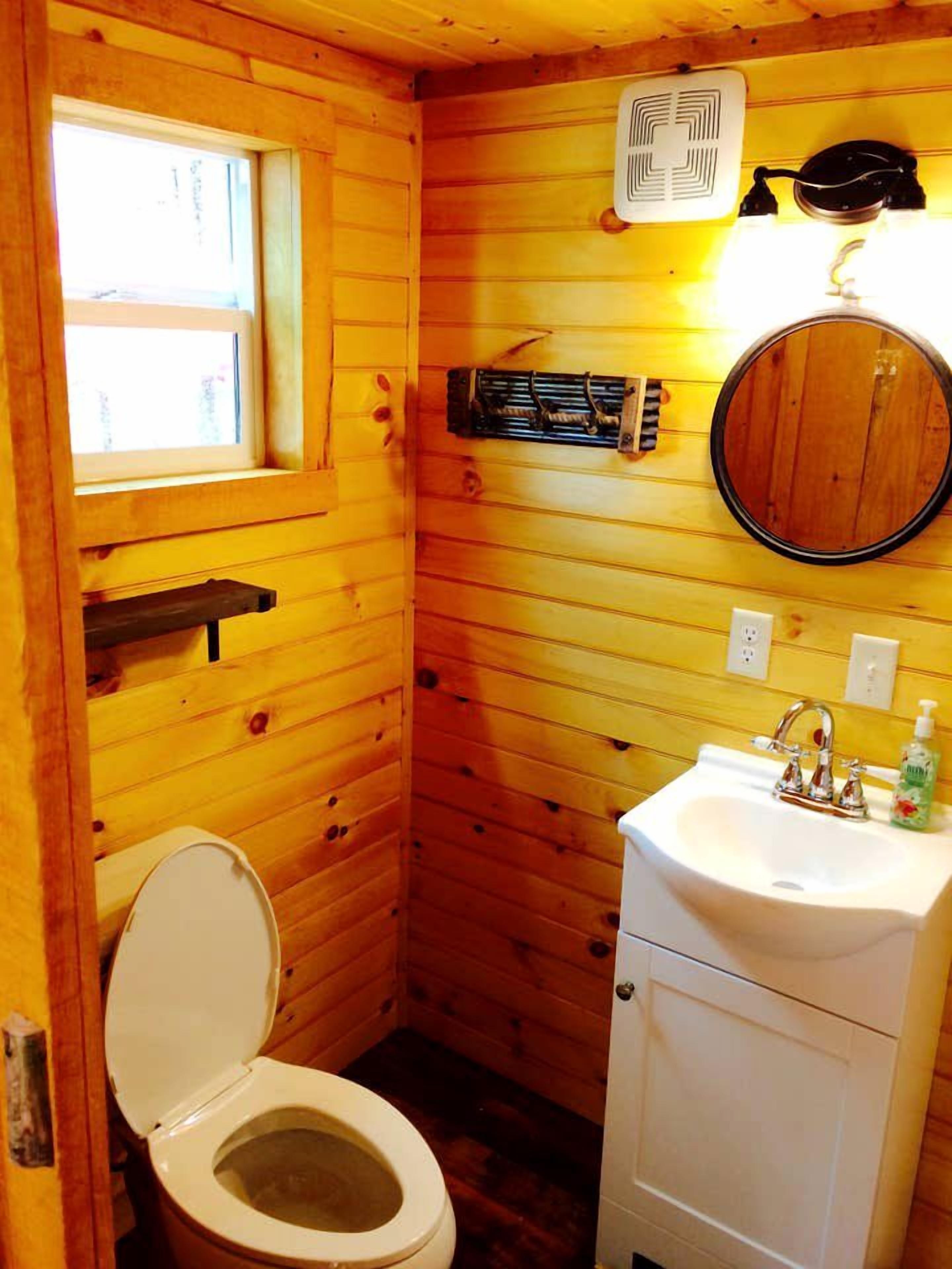 A wooden cabin bathroom with a toilet, sink, window, and a mirror.