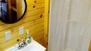A bathroom with a wooden wall, a white sink with a mirror, and a shower curtain. - Unique Wooden Tennessee Cabin near Knoxville for Relaxing Great Smoky Vacations (Newport)