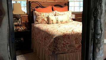 A cozy bedroom with a wooden ceiling, a bed with patterned bedding, a nightstand with a lamp, and a window with a view of trees.