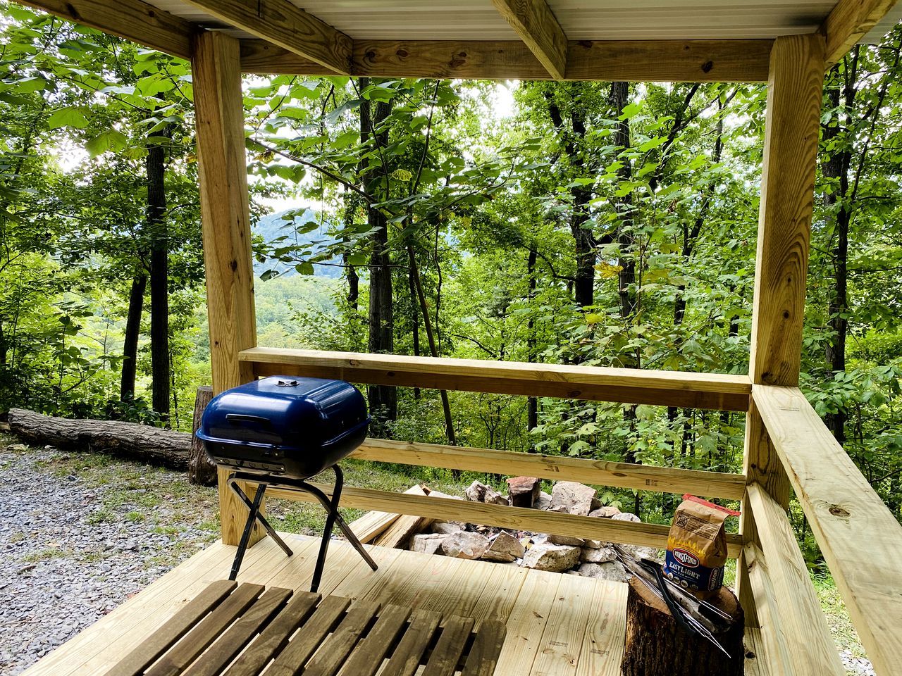 A wooden deck with a barbecue grill and firewood, surrounded by a forested area.