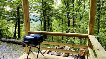 A wooden deck with a barbecue grill and firewood, surrounded by a forested area.