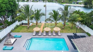 A swimming pool with lounge chairs and umbrellas, surrounded by palm trees and a view of a body of water. - Lakefront Tropical Villa by Glam Homes  (North Miami Beach)