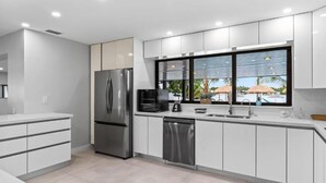 A modern kitchen with a stainless steel refrigerator, a dishwasher, and a window with a view of palm trees. - Lakefront Tropical Villa by Glam Homes  (North Miami Beach)