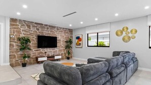 A modern living room with a stone accent wall, a flat-screen TV, a sectional sofa, and decorative wall art. - Lakefront Tropical Villa by Glam Homes  (North Miami Beach)