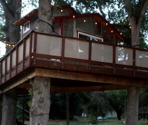 Ein Baumhaus mit einer Holzterrasse und einem kleinen, umzäumten Bereich, das/der von Bäumen getragen wird.