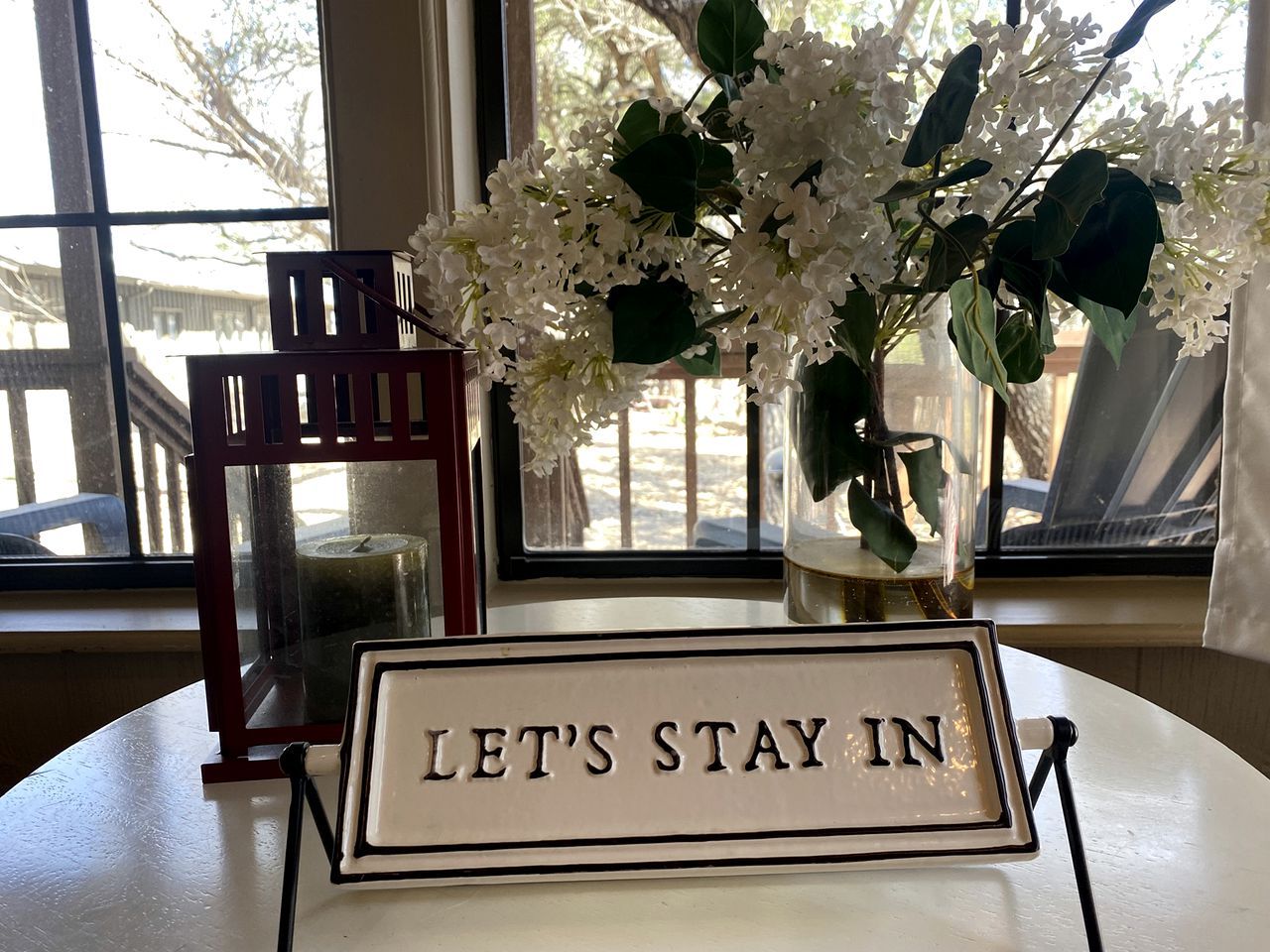 Una mesa con un cartel que dice 'LET'S STAY IN' y un florero con flores blancas.