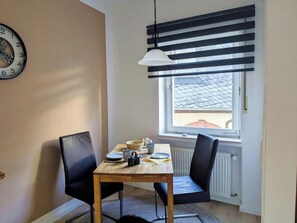 A dining area with a table, chairs, a clock, and a window with blinds. - Holiday apartment Brunnenplatz in our guesthouse in  old town of Zell (Moselle) (Zell (Mosel))