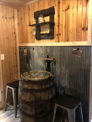 A rustic interior with wooden walls, a wooden barrel sink, stools, and a shelf with glasses. - Cozy Cabin Rental near Ozark National Forest for a Relaxing Weekend Getaway in Arkansas (Harrison)