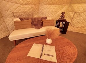 Une pièce chaleureuse avec un mur recouvert d’un tissu matelassé, un canapé blanc agrémenté de coussins marron, une petite table en bois sur laquelle repose un carnet, et un vase décoratif contenant des plumes.