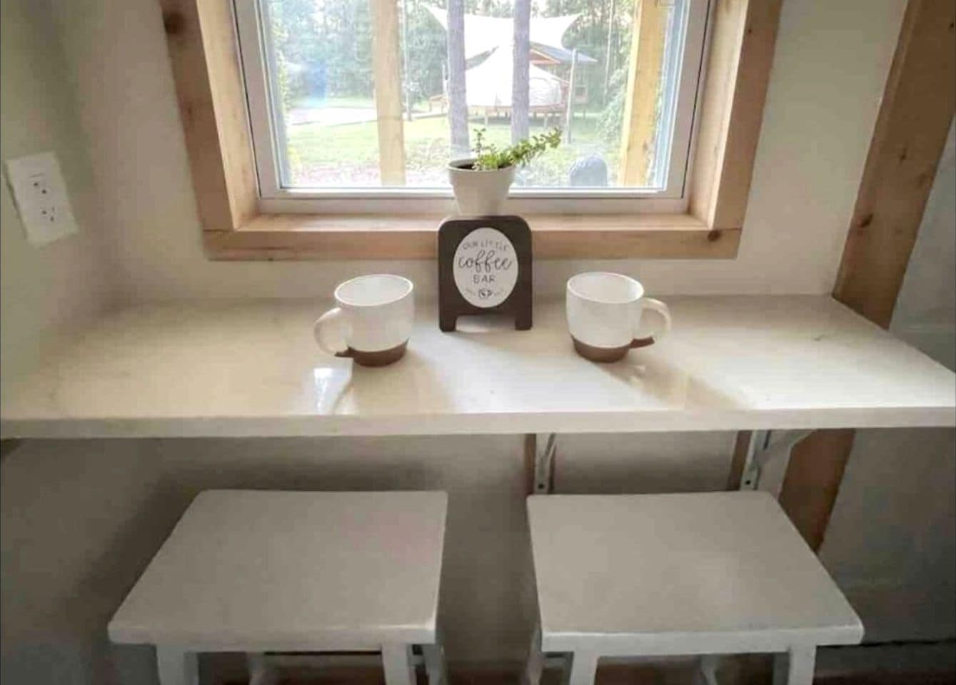 A small window with a view of trees and a building outside. Inside, there is a white shelf with two white mugs and a small potted plant. Below the shelf, there are two white tables.