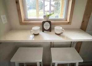 Une petite fenêtre donne sur des arbres et un bâtiment à l’extérieur. À l’intérieur, on aperçoit une étagère blanche sur laquelle sont posées deux tasses blanches et une petite plante en pot. En dessous de l’étagère, il y a deux petites tables blanches.