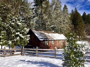 Una cabaña rústica con techo inclinado, rodeada de árboles cubiertos de nieve y una valla de madera.