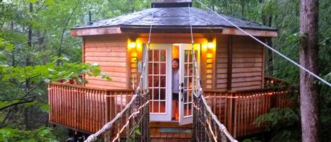 A wooden cabin with a rope bridge entrance, surrounded by trees.