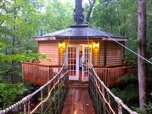 Una cabaña de madera con un puente colgante de acceso, rodeada de árboles.