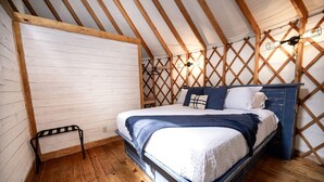 A bedroom with a wooden ceiling, a bed with white bedding, a wooden floor, and a small metal bench. - Family-Friendly Yurt with Hot-Tub near to the Medina River in Bandera, Texas (Bandera)