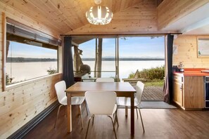 Uma cabana de madeira com mesa e cadeiras, cozinha e grandes janelas com vista para a água.