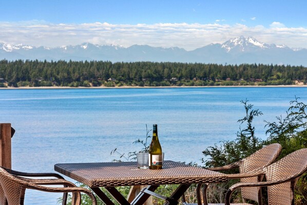 Une table en osier, avec une bouteille et un verre, donnant sur un lac bordé de montagnes.