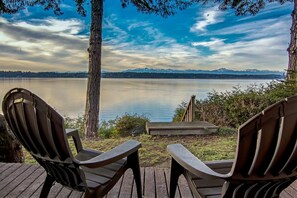 Two Adirondack chairs facing a lake with mountains in the distance, a wooden deck, and a bench.
