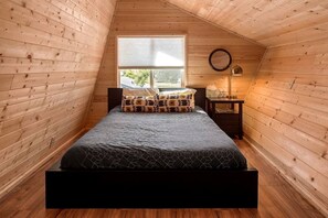 A bedroom with a wooden sloped ceiling, a bed with patterned bedding, a bedside table with a lamp, and a window with a view of greenery.