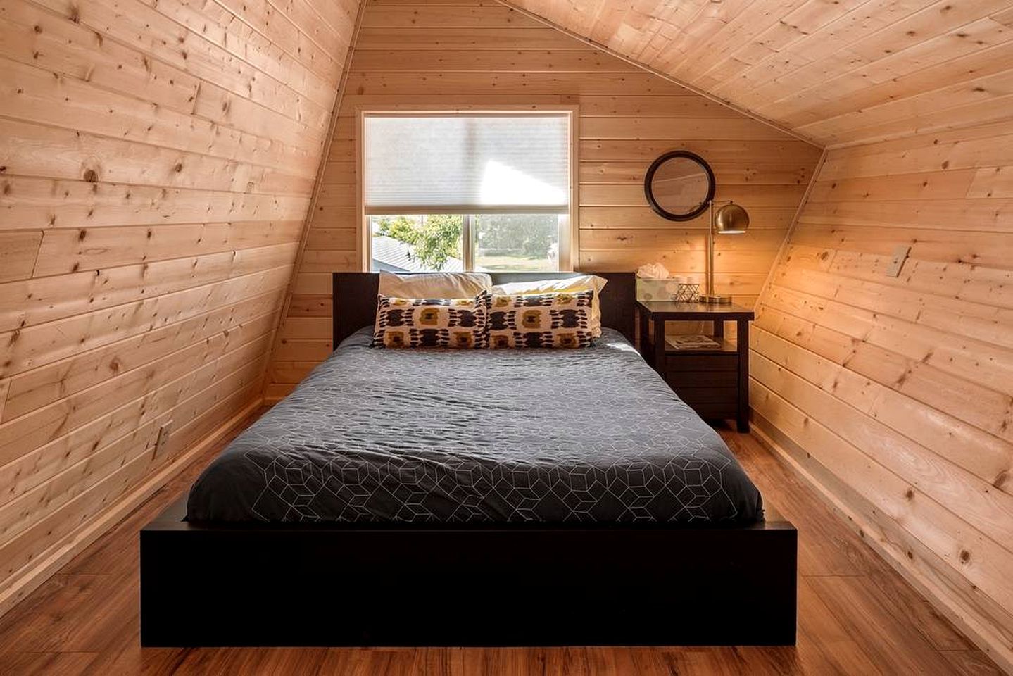 Un dormitorio con techo inclinado de madera, una cama con ropa de cama estampada, una mesita de noche con lámpara y una ventana con vistas a zonas verdes.
