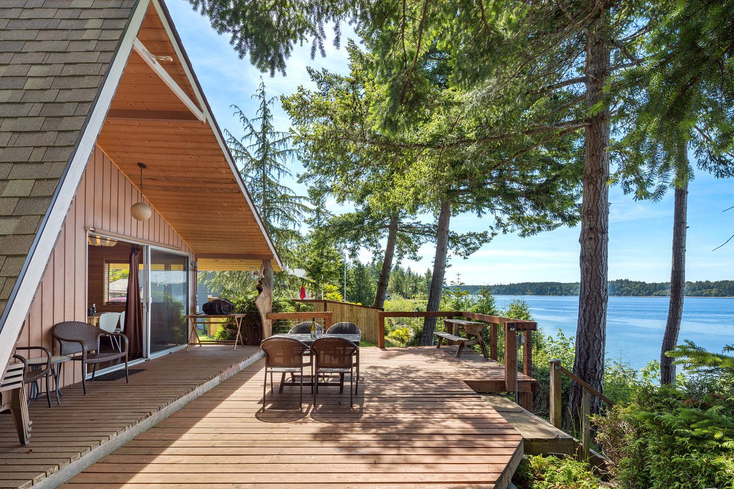 Une terrasse en bois avec des meubles d’extérieur, un espace couvert doté de portes-fenêtres coulissantes et une vue sur un lac entouré d’arbres.