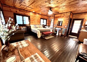 Ein Interieur einer Holzhütte mit einem Bett, einer Bank, einem Esstisch und einer Blumenvase.