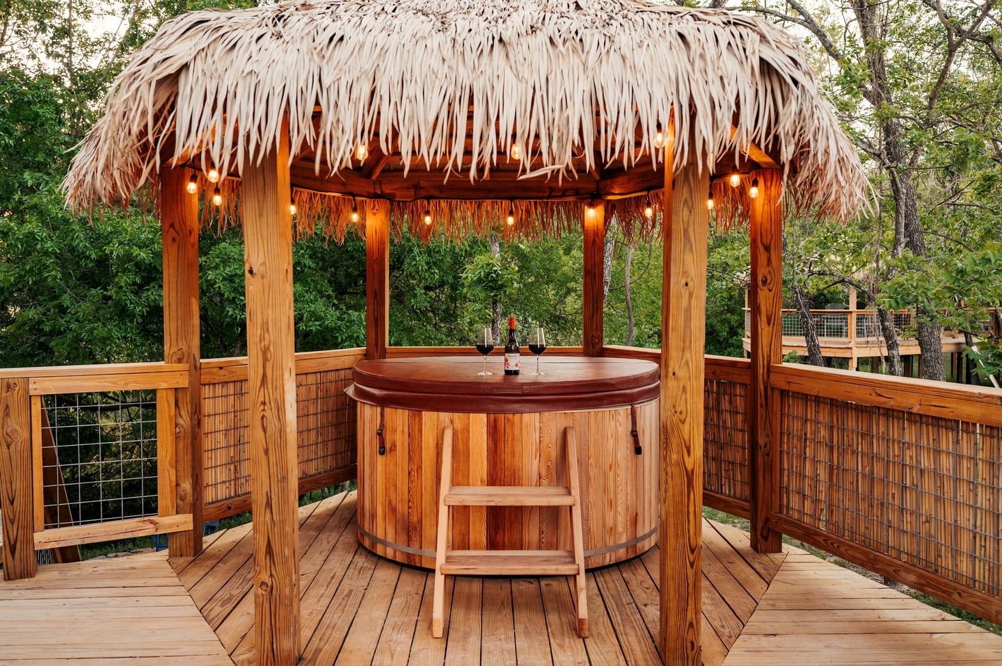 Un jacuzzi de madera con techo de paja, rodeado por una terraza de madera y una valla de bambú.
