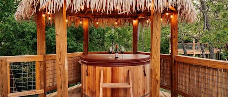 A wooden hot tub with a thatched roof, surrounded by a wooden deck and a bamboo fence.