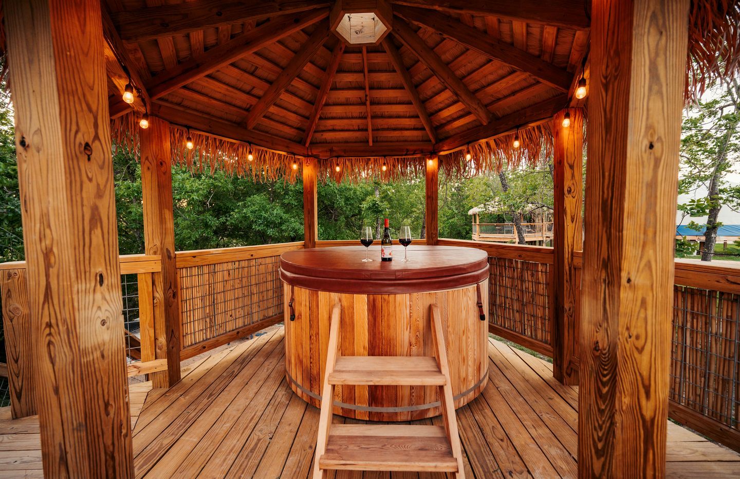A wooden gazebo with a hot tub, wooden deck, and a view of greenery.