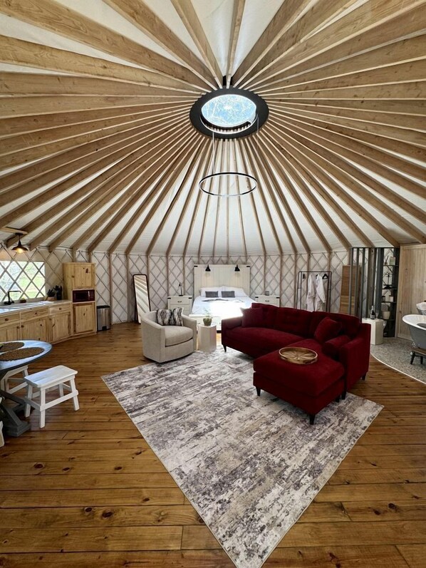 A room with a unique radial wooden ceiling design, a central circular light fixture, a bed with a headboard, a red sofa, a small round table, and a white chair. - Love Bird Yurt Romantic, Hot Tub, by the River in Bandera, Texas (Bandera)