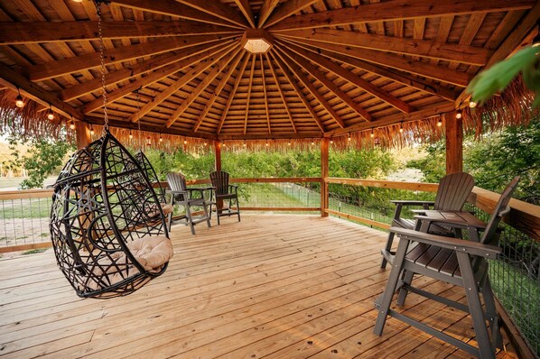 A wooden deck with a thatched roof gazebo, outdoor seating, and a hanging chair.