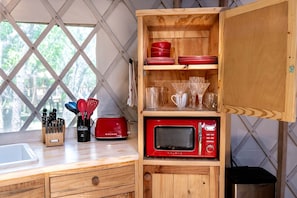 A compact kitchen with a microwave, a toaster, and a wooden cabinet with red dishes and glassware. - Love Bird Yurt Romantic, Hot Tub, by the River in Bandera, Texas (Bandera)