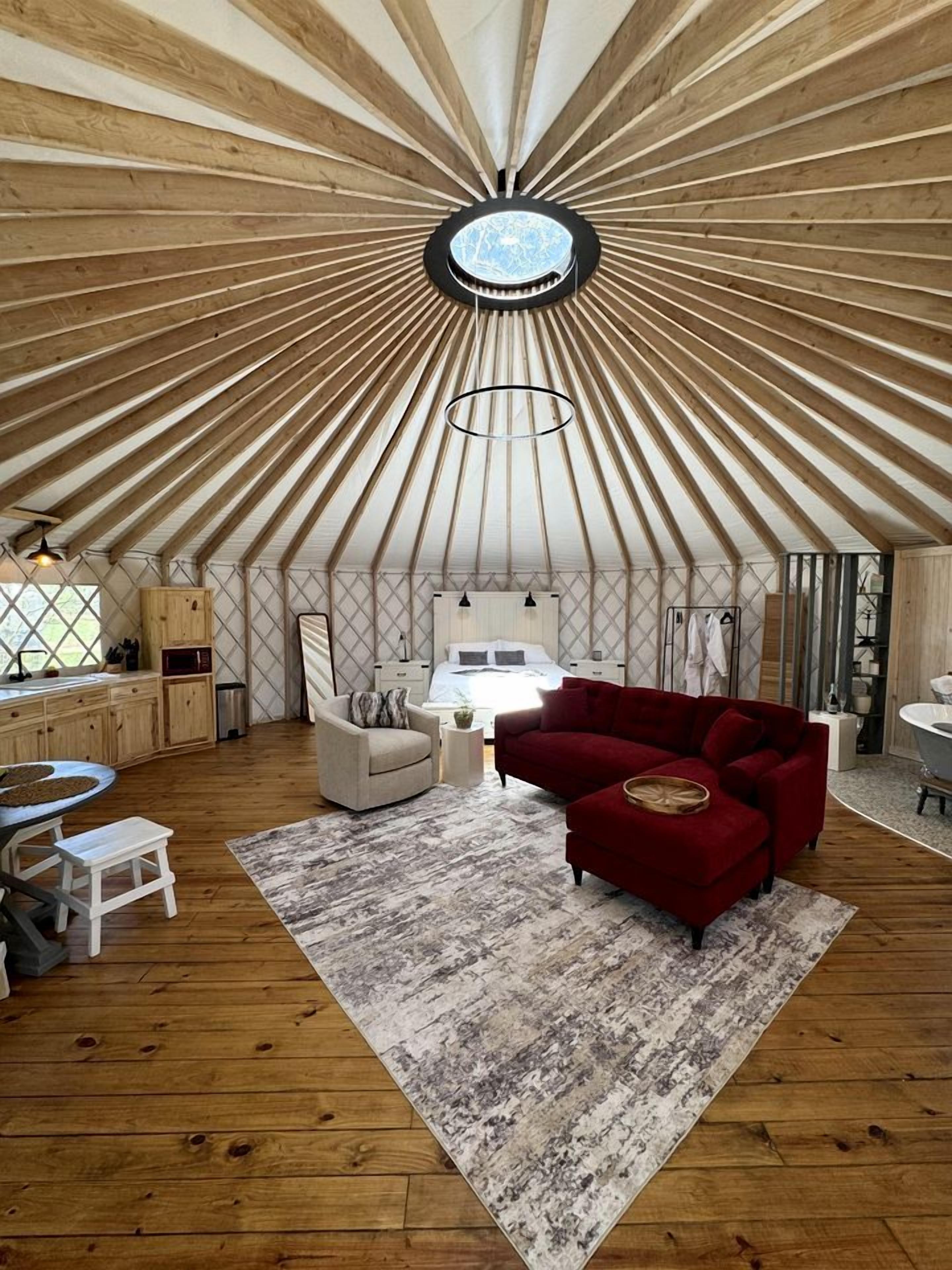 Una habitación con un diseño único de techo de madera radial, una lámpara circular central, una cama con cabecera, un sofá rojo, una pequeña mesa redonda y una silla blanca.