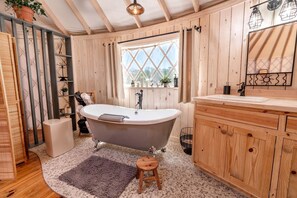 A bathroom with a freestanding bathtub, wooden cabinets, a window with a view, and a mirror with a sun design. - Love Bird Yurt Romantic, Hot Tub, by the River in Bandera, Texas (Bandera)