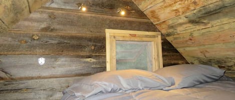 Una cabaña de madera con techo inclinado, una cama con colchón y almohadas grises, y un cuadro enmarcado en la pared.