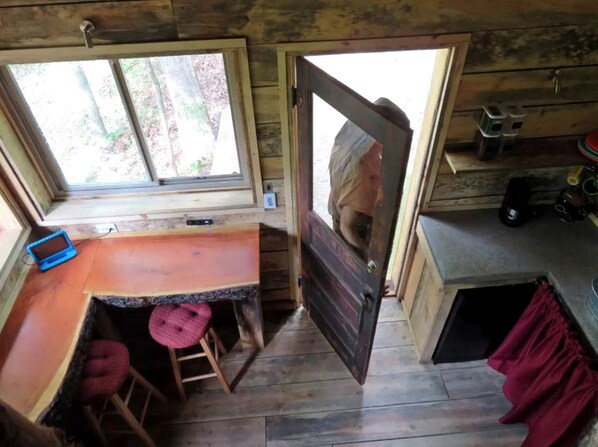 A cabin interior with a wooden dining table, a bench, a window, and a door leading outside.