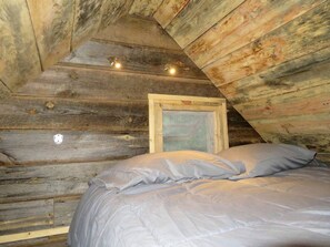 Een houten chalet met schuin dak, een bed met een grijze matras en kussens, en een lijst met een afbeelding aan de muur.