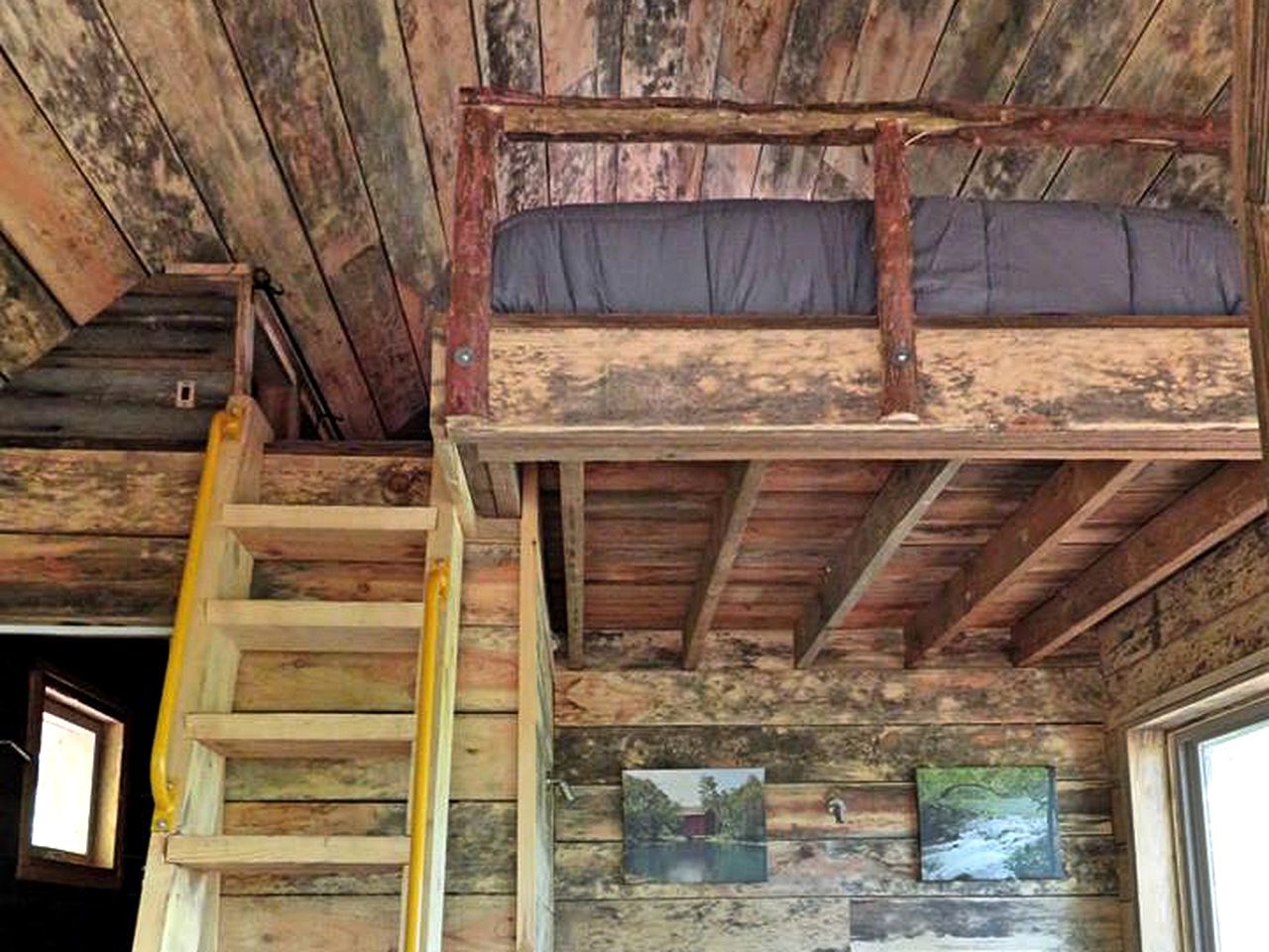 A rustic cabin interior with a wooden staircase, a loft bed, and framed pictures on the walls.