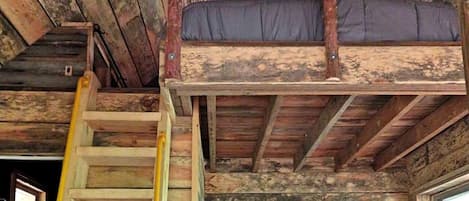 A rustic cabin interior with a wooden staircase, a loft bed, and framed pictures on the walls.